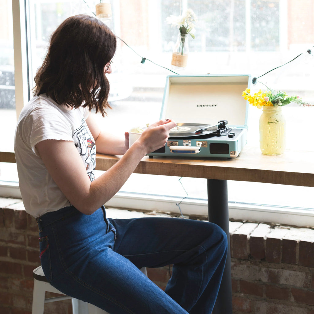 Woman playing vinyl record on blue Crosley turntable by a window with flowers in jars