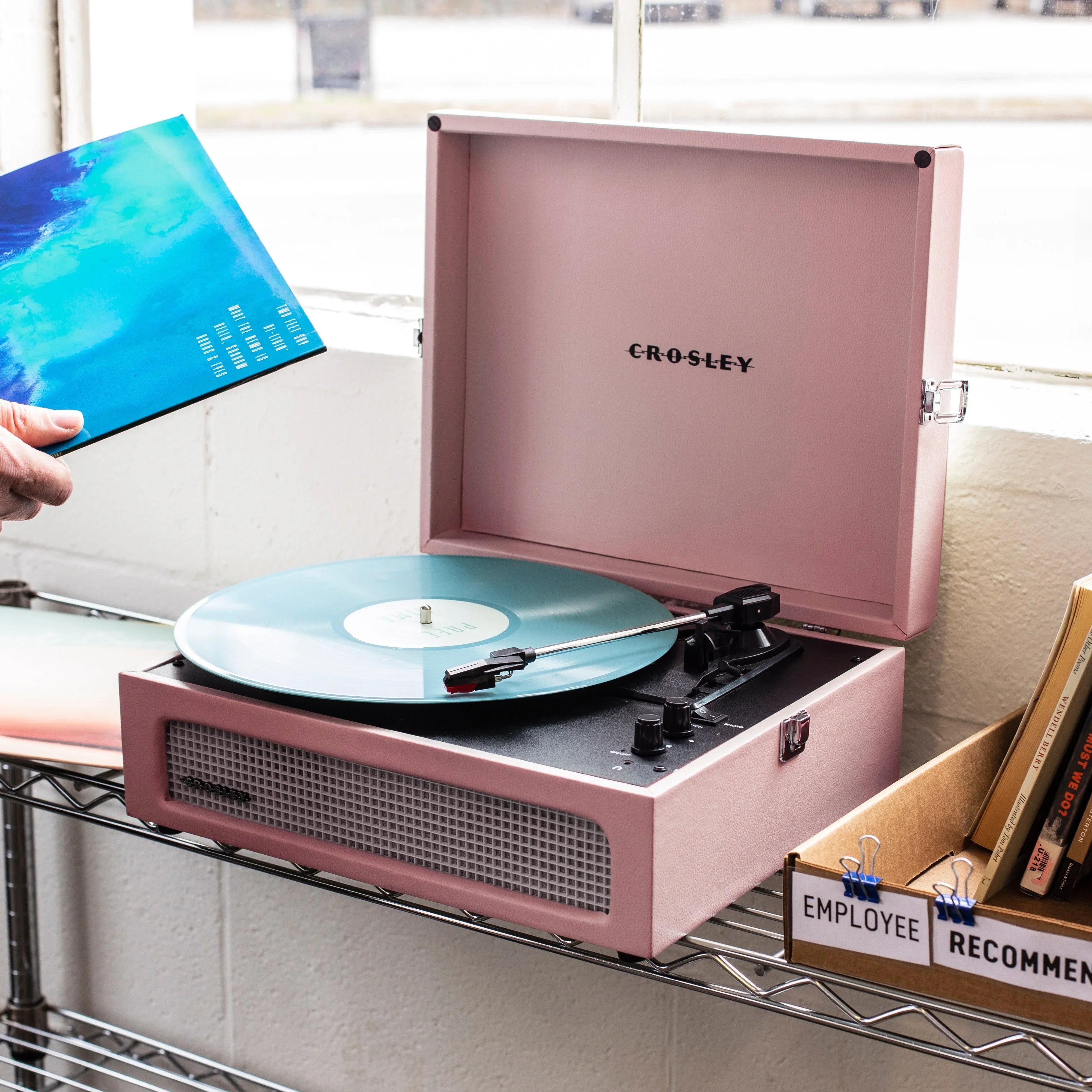 Pink Crosley record player with teal vinyl on metal shelf by window